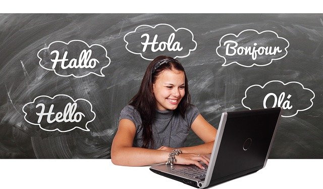 Eine junge Frau sitzt lächelnd vor ihrem Laptop und lernt Englisch. Im Hintergrund steht das Wort "Hallo" in verschiedenen Sprachen an einer Tafel.