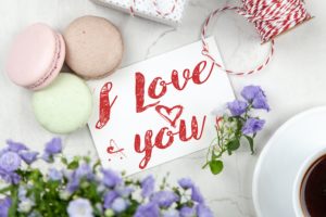 Auf einer Karte steht "I Love You" (Ich liebe dich). Neben der Karte liegen Kekse und es steht auch eine Kaffeetasse und ein Blumenstrauß daneben.