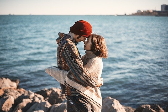 Ein junges Paar steht während der kalten Jahreszeit am Ufer des Meeres. Sie umarmen sich und schauen einander an.