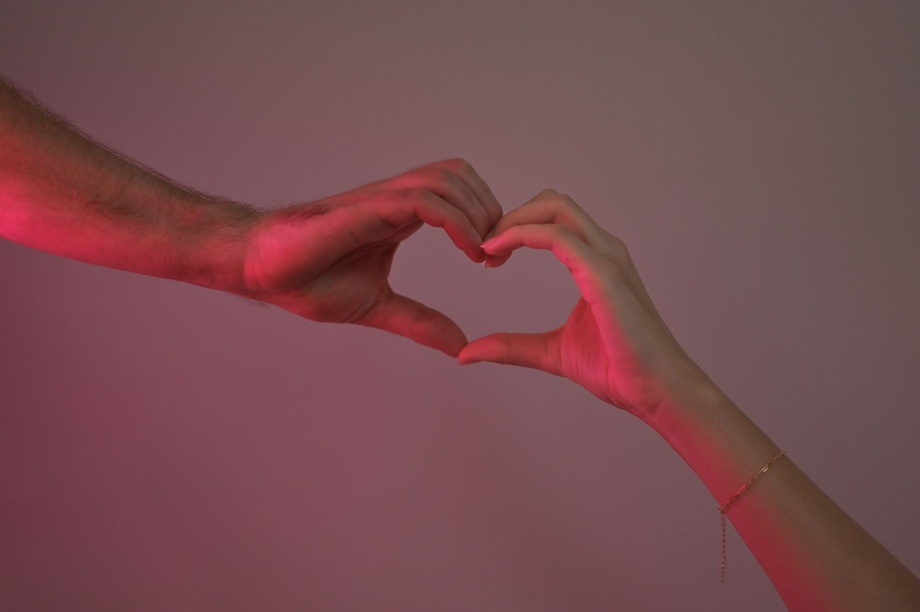 Two hands creating a heart shape against a pink backdrop, symbolizing love.