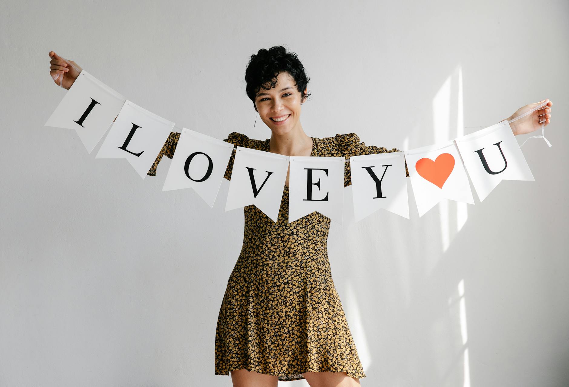 Smiling female looking at camera while standing on white background with decorative I Love You garland in hands during Saint Valentines day celebration